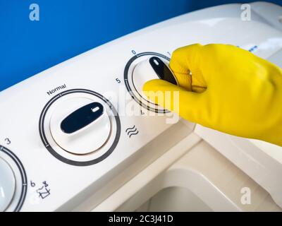 main d'une personne avec des gants jaunes commençant un blanc semi-automatique lave-linge avec fond bleu Banque D'Images