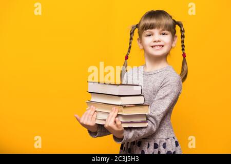 Photo de petite fille gaie avec des picots drôles tenant des livres d'école sur fond jaune. Retour à l'école Banque D'Images