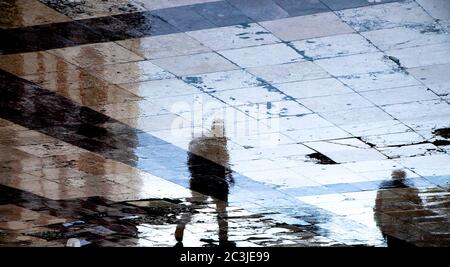 Silhouette floue et reflet ombre des personnes marchant sur une flaque de rue humide dans la zone piétonne de la ville par temps pluvieux Banque D'Images