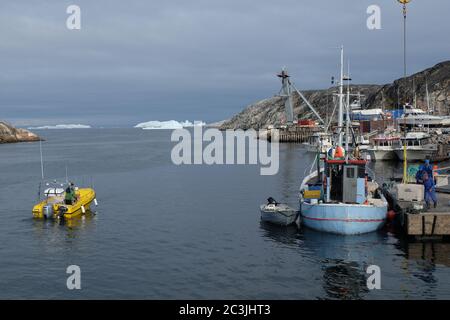 Un petit bateau de pêche Greenlandic décharge sa prise dans le port d'Iluissat tandis qu'un chasseur de phoques se dirige vers les flotteurs de glace. Banque D'Images