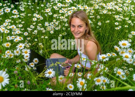 Une belle jeune fille avec un ordinateur portable est assise dans un pré parmi les pâquerettes fleuries et souriant doux. Banque D'Images