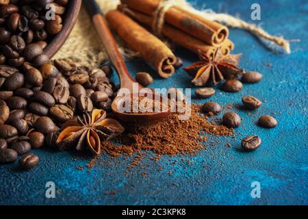 grains de café dispersés sur un fond texturé bleu, étoiles anis, bâtonnets de cannelle et café moulu dans une cuillère en bois. Banque D'Images