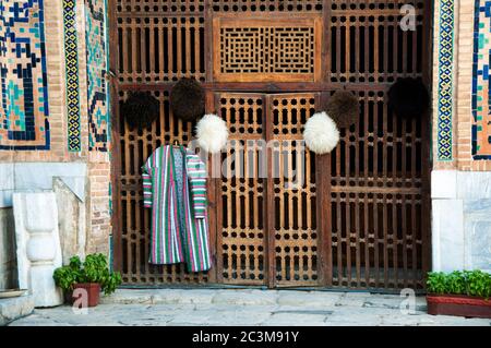 Porte en bois décorative dans la cour intérieure du Registan avec des chapeaux de fourrure et une robe traditionnelle ouzbek colorée manteau accroché à une porte, Samarkand, Ouzbékistan Banque D'Images