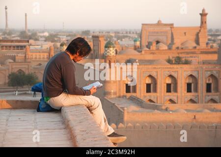 Un touriste solitaire assis sur un haut de l'ancien mur, lisant un livre pendant le beau coucher de soleil d'or sur l'ancienne ville de Khiva, Ouzbékistan Banque D'Images