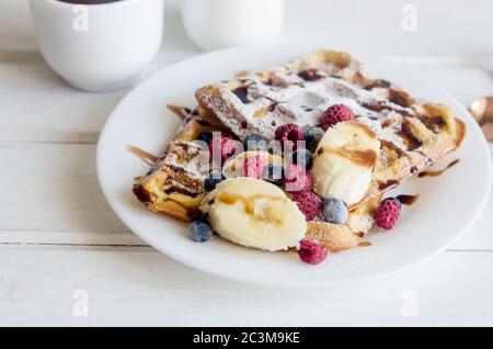 Assiette blanche de gaufres belges avec sucre en poudre, baies glacées, sauce banane et caramel avec une tasse de latte de café sur une table en bois blanc, pause savoureuse Banque D'Images