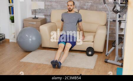 Un homme fort qui fait de l'entraînement triceps en utilisant le canapé dans le salon portant des vêtements de sport. Banque D'Images