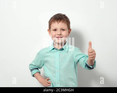 Un garçon de quatre ans sourit et montre le pouce vers le haut. Enfant heureux en chemise bleue, isolée sur fond blanc. Banque D'Images