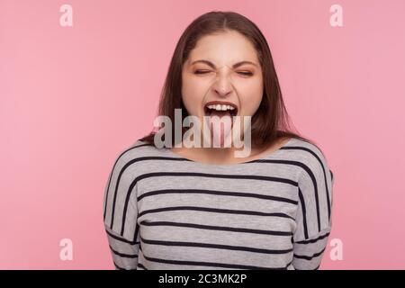 Portrait de femme drôle désobéissante en sweat-shirt rayé montrant la langue, exprimant le mécontentement, taquinant avec le grimace délice, beh méchant Banque D'Images