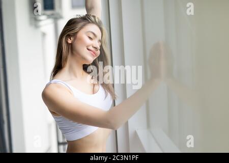 Jeune femme souriante en robe de nuit en dentelle se relaxant dans une maison confortable, se sentant heureuse, se reposant le matin, regardant par la fenêtre Banque D'Images