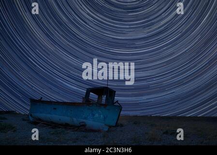 Image numérique composite des pistes étoiles autour de Polaris avec un bateau de pêche abandonné sur des paysages de plage de galets Banque D'Images