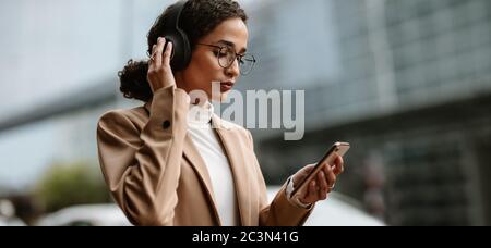 Femme d'affaires écoutant de la musique sur son téléphone mobile. Femme d'affaires portant un casque et regardant son téléphone cellulaire dans la rue. Banque D'Images