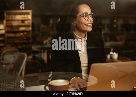 Femme d'affaires dans un café avec ordinateur portable donnant à l'extérieur de la fenêtre et souriant. Femme assise au café avec son ordinateur portable. Banque D'Images
