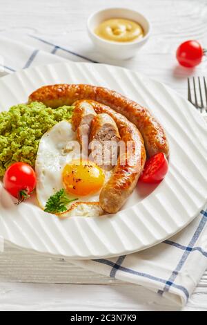 Assiette blanche avec de longues saucisses de porc, œufs frits côté ensoleillé, tomates cerises, purée de pois verts sur un fond en bois clair avec moutarde et couverts Banque D'Images