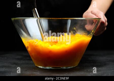 Femme cuisant de la purée de citrouille avec un mélangeur dans un bol en verre transparent sur une table noire. Purée de citrouille. Aliments sains à base de légumes Banque D'Images