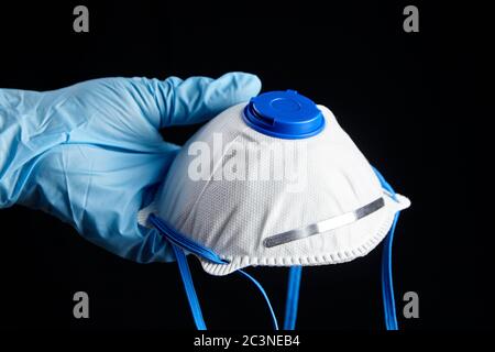 Masque FFP2 en main dans un gant en caoutchouc bleu, isolé sur fond noir. Protection contre la pandémie de coronovirus COVID-19. Masque filtrant blanc Banque D'Images