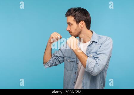 Vue latérale d'un homme en maillot denim ouvrier debout avec geste de boxe, poinçonnement à la caméra, provocature de combat ou prêt à se défendre, combat concept. i Banque D'Images