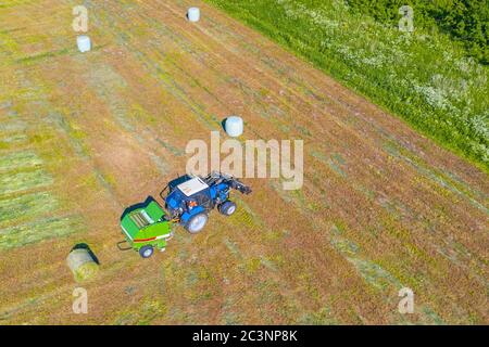 Un tracteur agricole recueille l'herbe fauchée pour une utilisation agricole et encapsule les balles de foin dans un champ en plastique, vue aérienne du dessus Banque D'Images