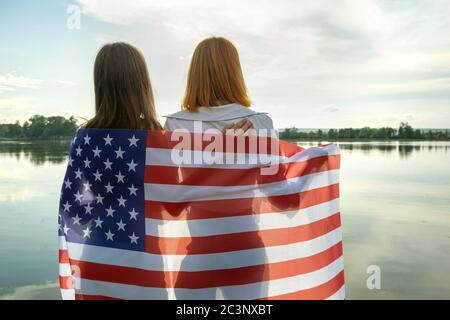 Deux jeunes amies femmes avec drapeau national des États-Unis sur leurs épaules se serrent à l'extérieur sur le bord du lac. Jeunes filles patriotiques célébrant les États-Unis i Banque D'Images