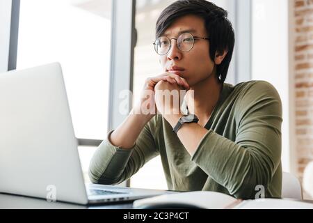 Image d'un jeune asiatique élégant portant des lunettes à l'aide d'un ordinateur portable assis à une table dans l'appartement Banque D'Images