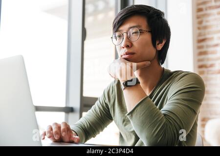 Image d'un jeune asiatique élégant portant des lunettes à l'aide d'un ordinateur portable assis à une table dans l'appartement Banque D'Images