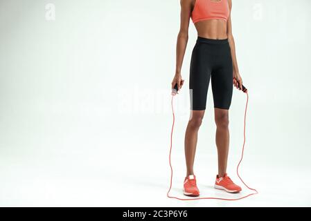 Mode de vie actif. Photo courte d'une jeune femme africaine avec un corps parfait dans des vêtements de sport corde à sauter tout en faisant de l'exercice en studio sur fond gris. Équipement de sport. Concept de fitness Banque D'Images