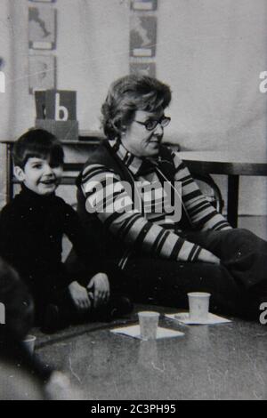 Belle photo vintage des années 70 en noir et blanc d'un garçon assis sur le sol en s'amusant avec son professeur. Banque D'Images
