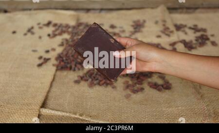 Femme tenant une main d'un bloc de chocolat fait à la main avec des fèves de cacao sèches sur une table couverte de jute dans le fond Banque D'Images