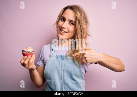 Jeune belle femme blonde eatimg chocolat cupcake sur fond rose isolé avec le visage surprise pointant du doigt vers lui-même Banque D'Images