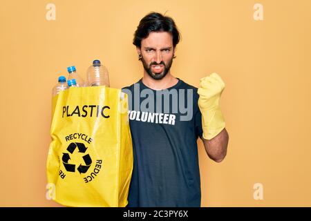 Beau volontaire hispanique portant le t-shirt de bénévolat sac de maintien avec des bouteilles en plastique agacé et frustré criant avec colère, fou et vous Banque D'Images