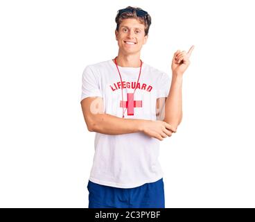 Jeune homme beau portant le maître-nageur t-shirt et siffler souriant heureux pointant avec la main et le doigt sur le côté Banque D'Images