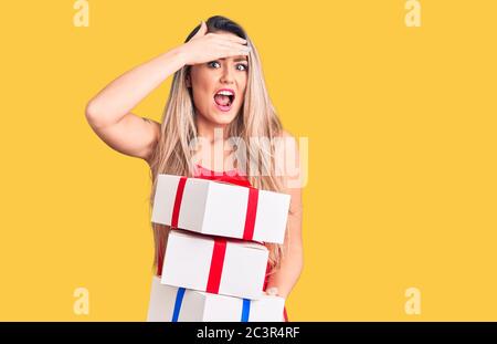 Jeune belle femme blonde tenant des cadeaux d'anniversaire stressée et frustrée avec la main sur la tête, surprise et colère visage Banque D'Images