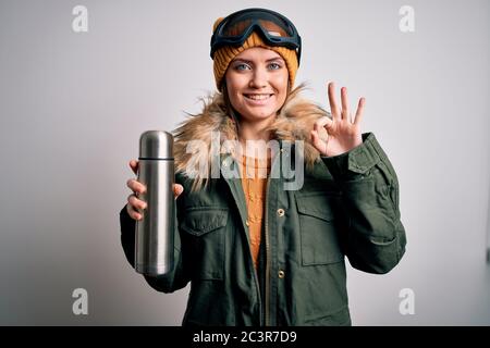 Jeune belle skieuse femme avec les yeux bleus portant des lunettes de ski buvant thermo avec café faisant signe ok avec les doigts, excellent symbole Banque D'Images