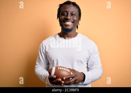 Jeune joueur africain jouant au rugby tenant le ballon de football américain sur fond jaune avec un visage heureux debout et souriant avec un s confiant Banque D'Images