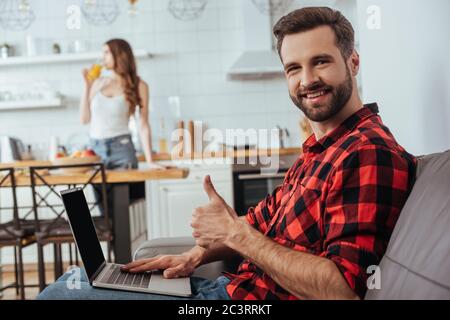 un freelance souriant montrant le pouce tout en travaillant sur un ordinateur portable dans la cuisine, près de la petite amie en arrière-plan Banque D'Images