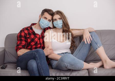 jeune couple déprimé dans des masques médicaux assis sur un canapé et regardant l'appareil photo Banque D'Images