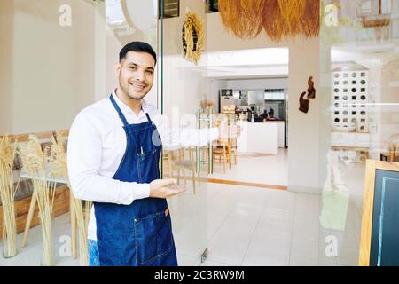 Beau propriétaire souriant de boulangerie dans le tablier faisant le geste de bienvenue et invitant les clients à l'intérieur Banque D'Images