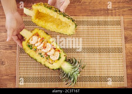 Femme mettant du riz frit avec des légumes et des crevettes dans un bol d'ananas, vue d'en haut Banque D'Images