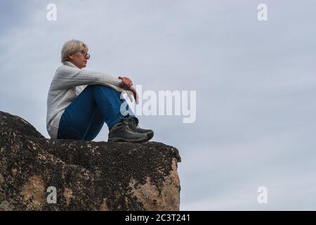 Femme couvée avec des lunettes, vêtue d'un chandail blanc et d'un Jean. Il est situé au sommet d'une montagne contre le ciel et les nuages. Concept de voyage. Banque D'Images