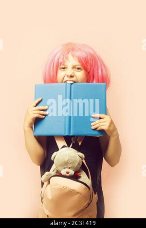 Fille avec sac à dos et jouet morsures grand livre bleu. Écolière avec perruque rose isolée sur fond rose clair. Enfant avec un visage en colère en uniforme scolaire. Concept de l'enfance et de la rentrée. Banque D'Images