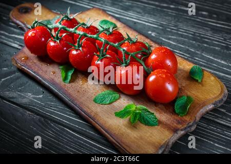 Tomates sur une branche. Tomates sur une belle branche verte dans des gouttes d'eau avec des feuilles de menthe sur un plan de cuisine en bois. Banque D'Images