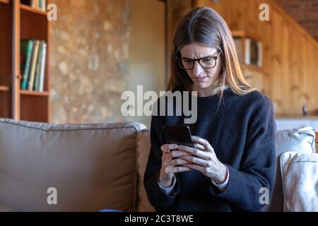 Jeune femme brune assez sérieuse avec des lunettes dactylographiant sur le smartphone assis sur le canapé à la maison portant un chandail noir Banque D'Images