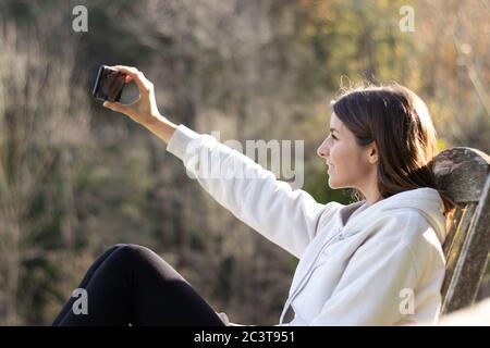 Belle femme souriante brune prend un selfie avec le smartphone assis dans le parc lors d'une journée ensoleillée. Banque D'Images