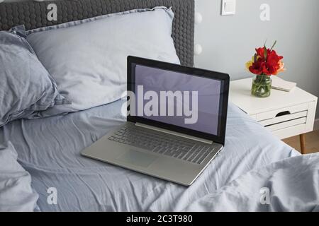 Lit vide gris embrouillé avec ordinateur portable dans une chambre minimaliste avec fleurs sur la table de nuit Banque D'Images
