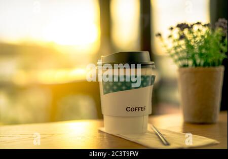 Café en papier dans le café sur des fleurs floues avec des feuilles vertes dans la casserole sur une table en bois brun dans le café le matin avec la lumière du soleil. Banque D'Images