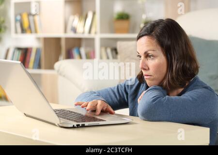 Femme adulte sérieuse vérifiant le contenu en ligne sur ordinateur portable assis sur le sol dans le salon à la maison Banque D'Images