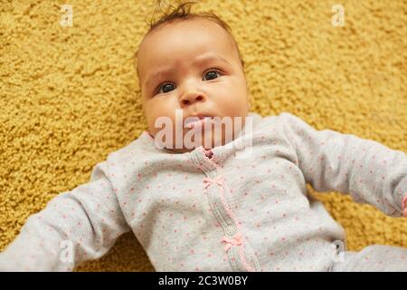 Au-dessus de la vue portrait de bébé de course mixte mignon regardant l'appareil photo tout en étant allongé sur la moquette jaune moelleuse, espace de copie Banque D'Images