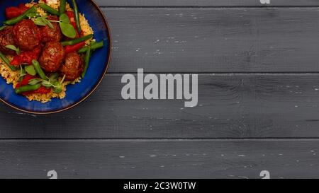 Un délicieux bol de boulettes de viande chipolte, couscous et légumes, dans un bol bleu rustique sur fond de bois foncé en format bannière Banque D'Images