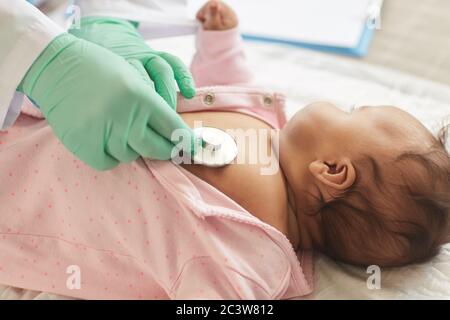 Gros plan d'un médecin méconnaissable utilisant un stéthoscope tout en examinant un bébé mixte mignon allongé sur une table à langer en clinique, espace de copie Banque D'Images