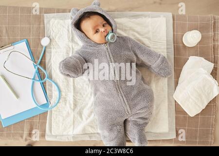 Au-dessus de la vue sur un adorable bébé afro-américain portant une combinaison en peluche, allongé sur une table à langer pendant le contrôle au bureau du médecin, espace copie Banque D'Images