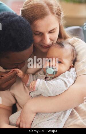 Portrait vertical à tons chauds de la famille interraciale aimante tenant le bébé garçon, avec un père afro-américain embrassant doucement la petite main Banque D'Images